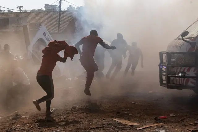 Un nuage de gaz lacrymogène dessine la silhouette d'un groupe de personnes qui tentent de courir sur un sol poussiéreux pour échapper à la fumée.
