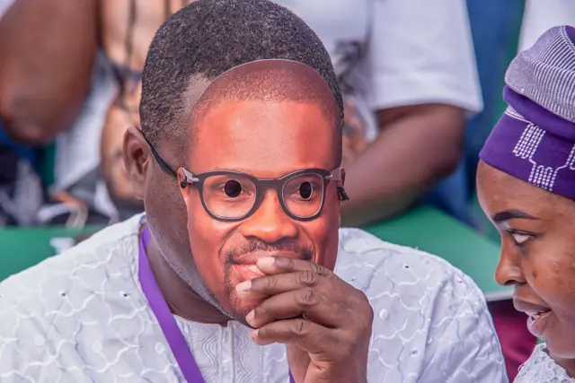 Un homme vêtu d'une chemise grise tient devant son visage un masque à l'effigie du ministre des Finances du Bénin. 