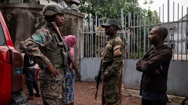 Deux militaires en uniforme, fusil à l’épaule, accompagnés d’un civil en sweat à capuche marron et d’une femme en sweat rose en arrière-plan
